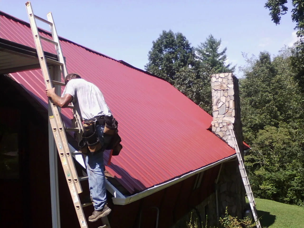Roofer on a ladder beside a red metal roof and stone chimney — Soffit & Fascia Repair by Roofing Brothers in Lake Peekskill
